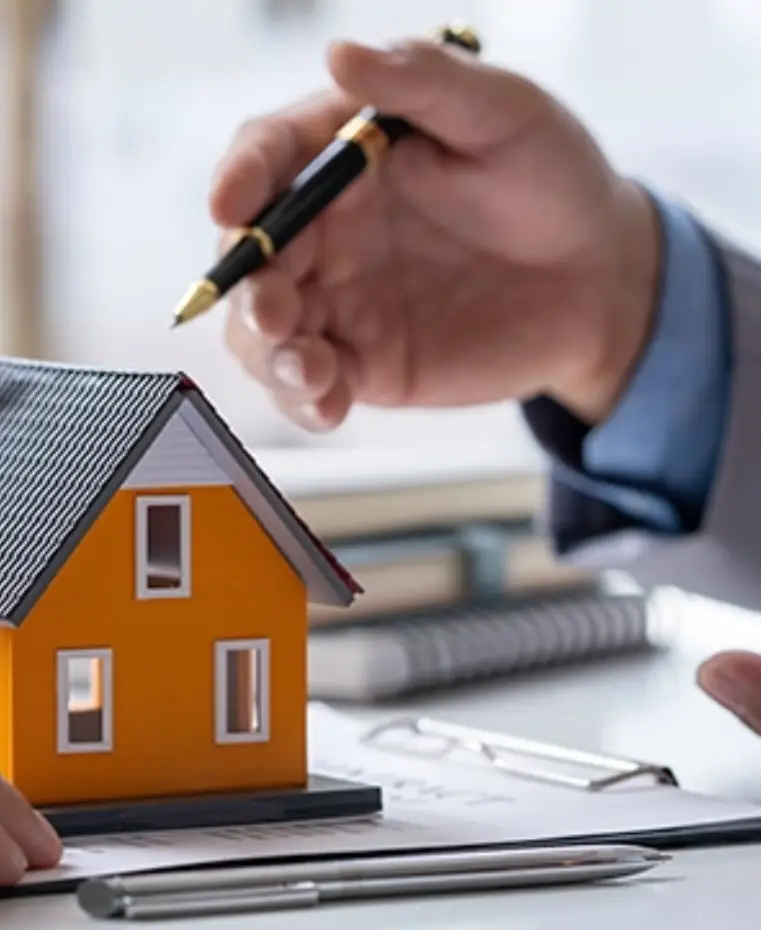Two hands gesturing over a small orange model house on a desk, with pens and papers visible, suggesting a discussion about real estate
